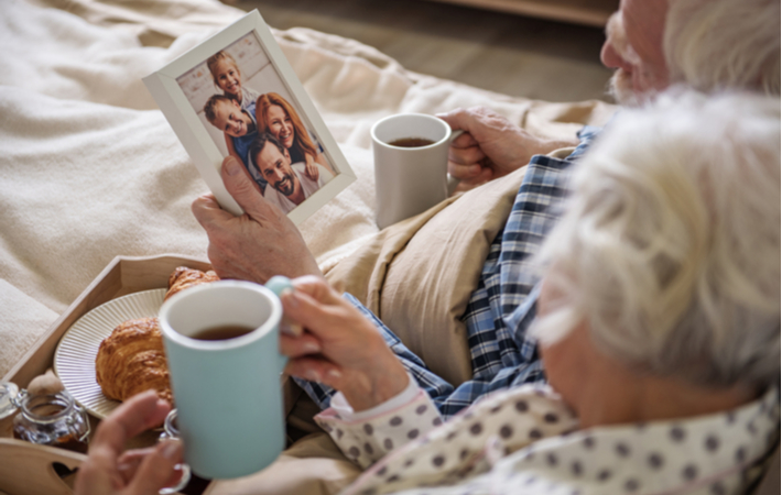 Senior couple looking at family picture while laying in bed with coffee and snacks at senior community memory care