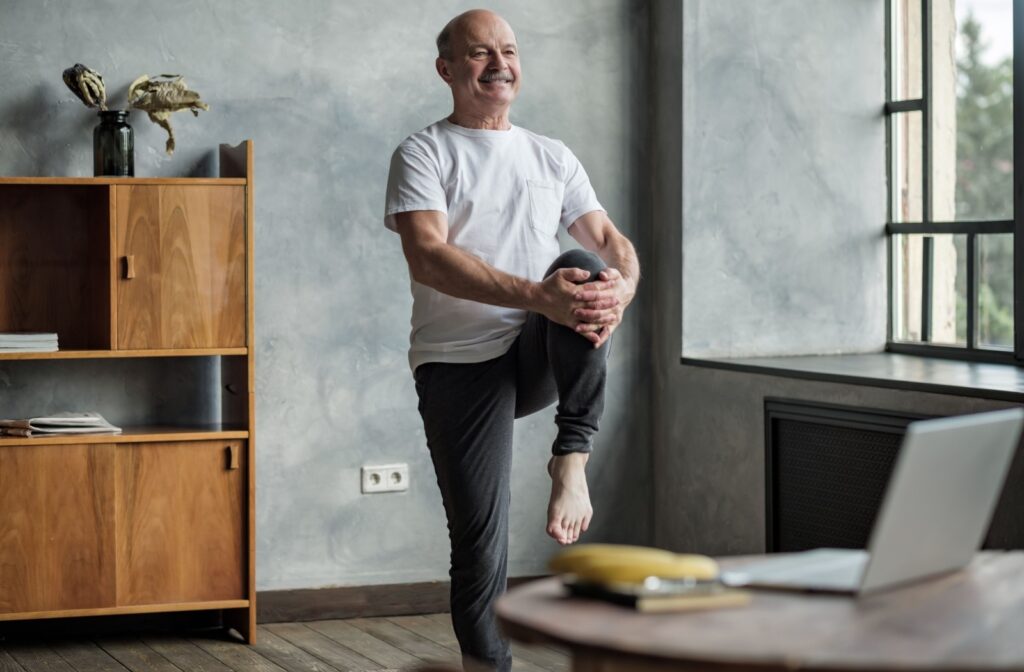 A smiling senior performs a single-leg balance exercise from the comfort of their home in a senior living community.