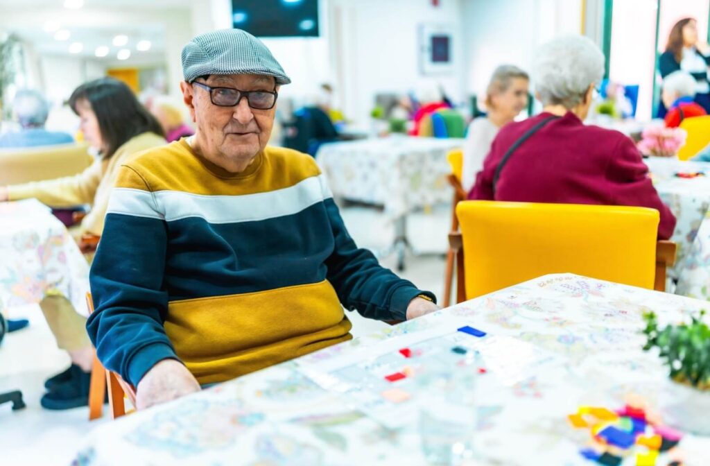 An older adult smiles and laughs while playing a small plastic block game in a crowded common area of a senior living community