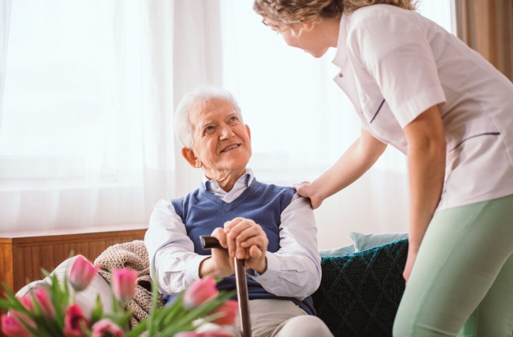 An older adult sits in front of a window and smiles up at a caregiver during a check-in
